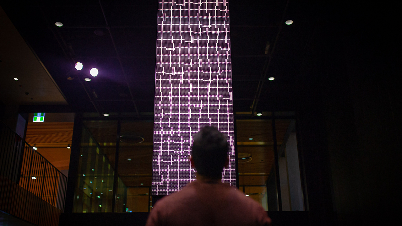 Man stars upward at purple flip dot screen.