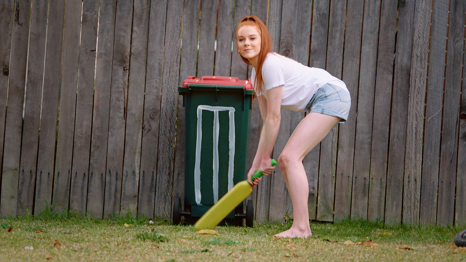 Woman with cricket bat assuming batting stance in front of stumps drawn on a garbage bin.